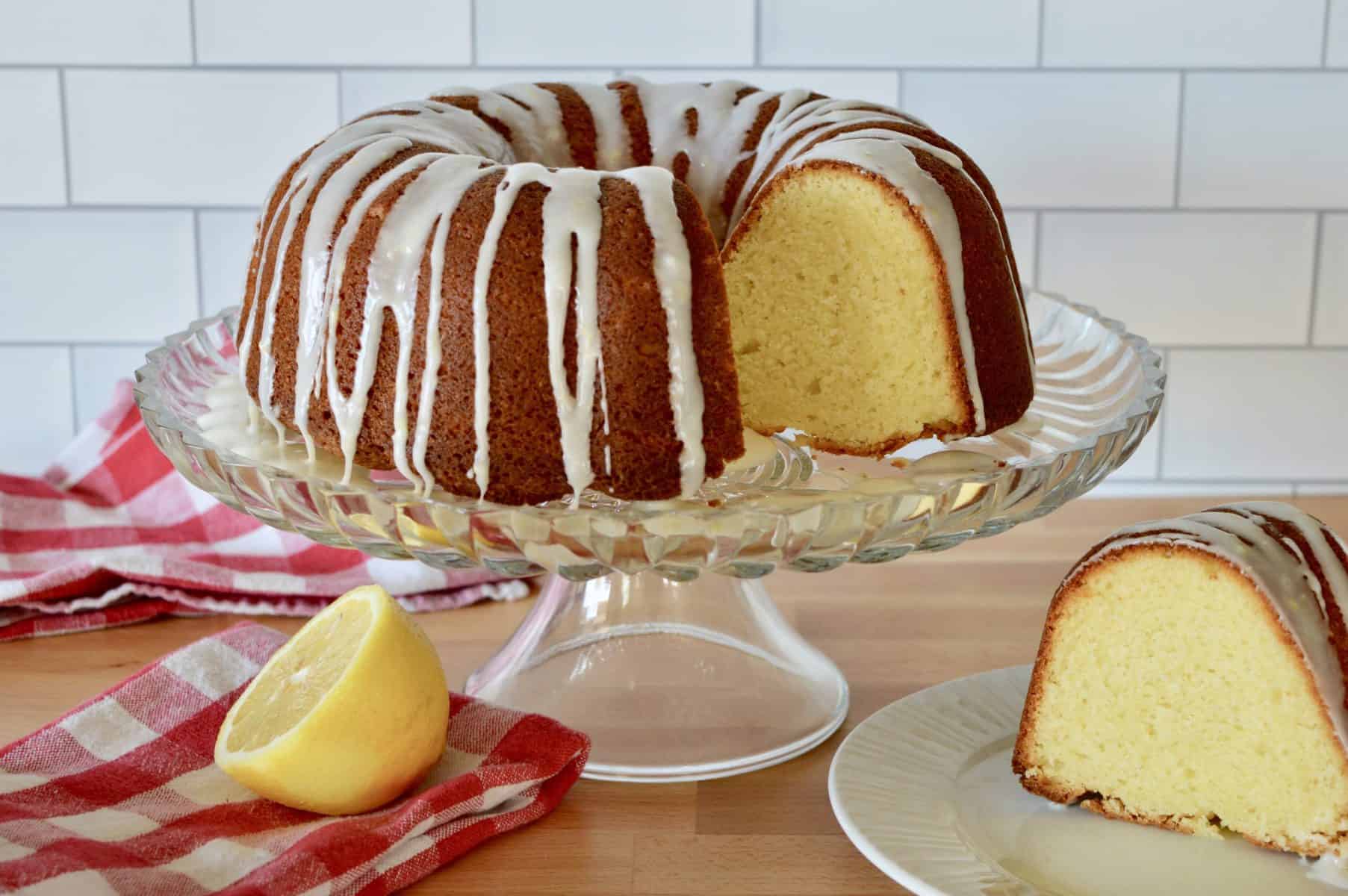 Olive Oil Bundt Cake on a pedestal with a slice of cake on a white plate and a half of a lemon. 