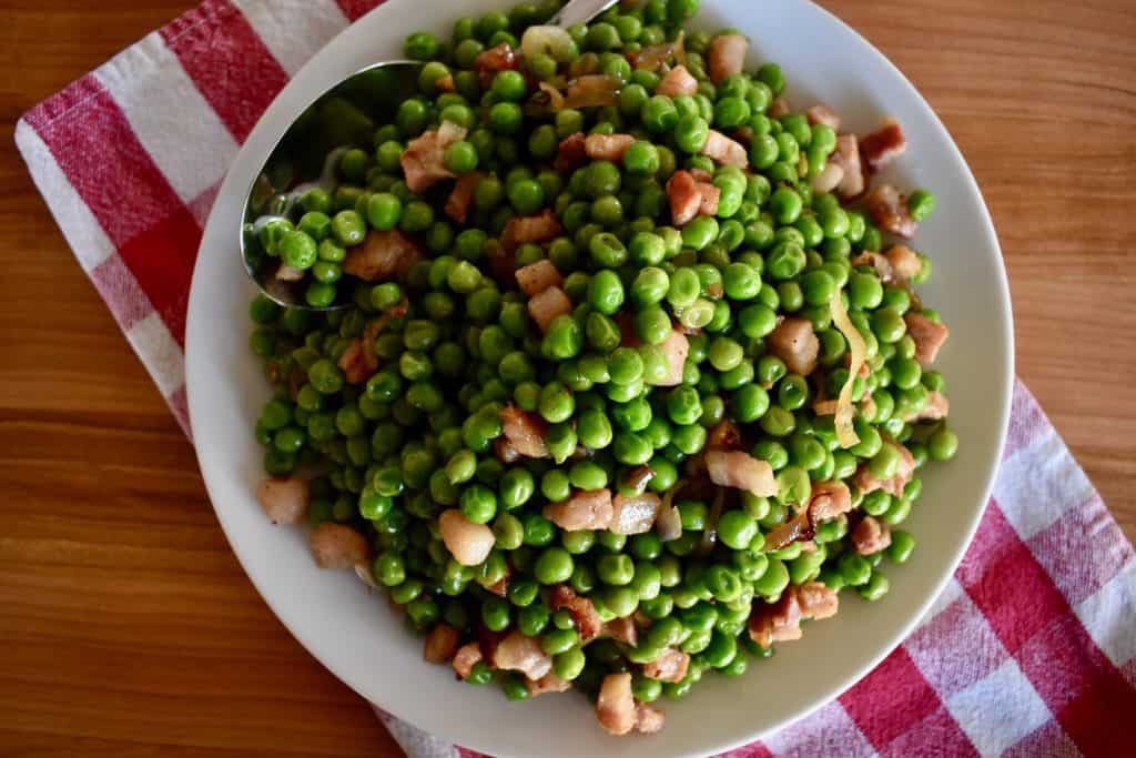 Overhead photo of peas and pancetta in a white dish with a checkered towel underneath.