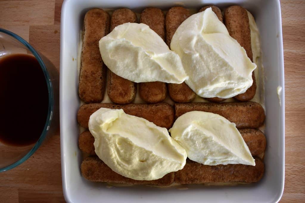 Scoops of mascarpone custard on the ladyfingers layer in a white dish.