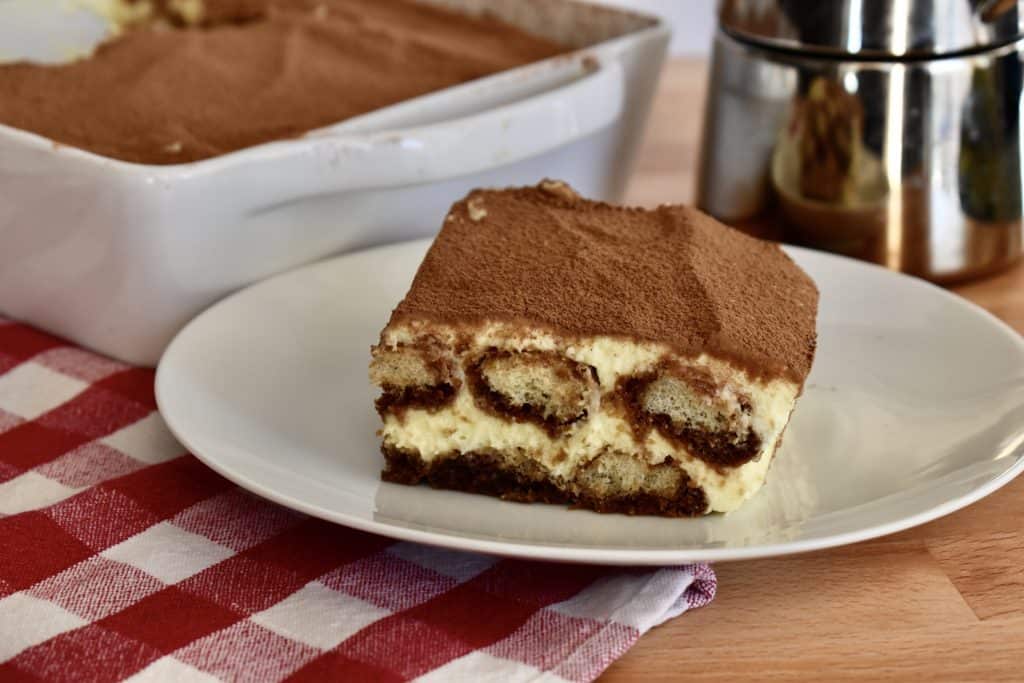 Tiramisu on a white plate with an espresso maker in the background.