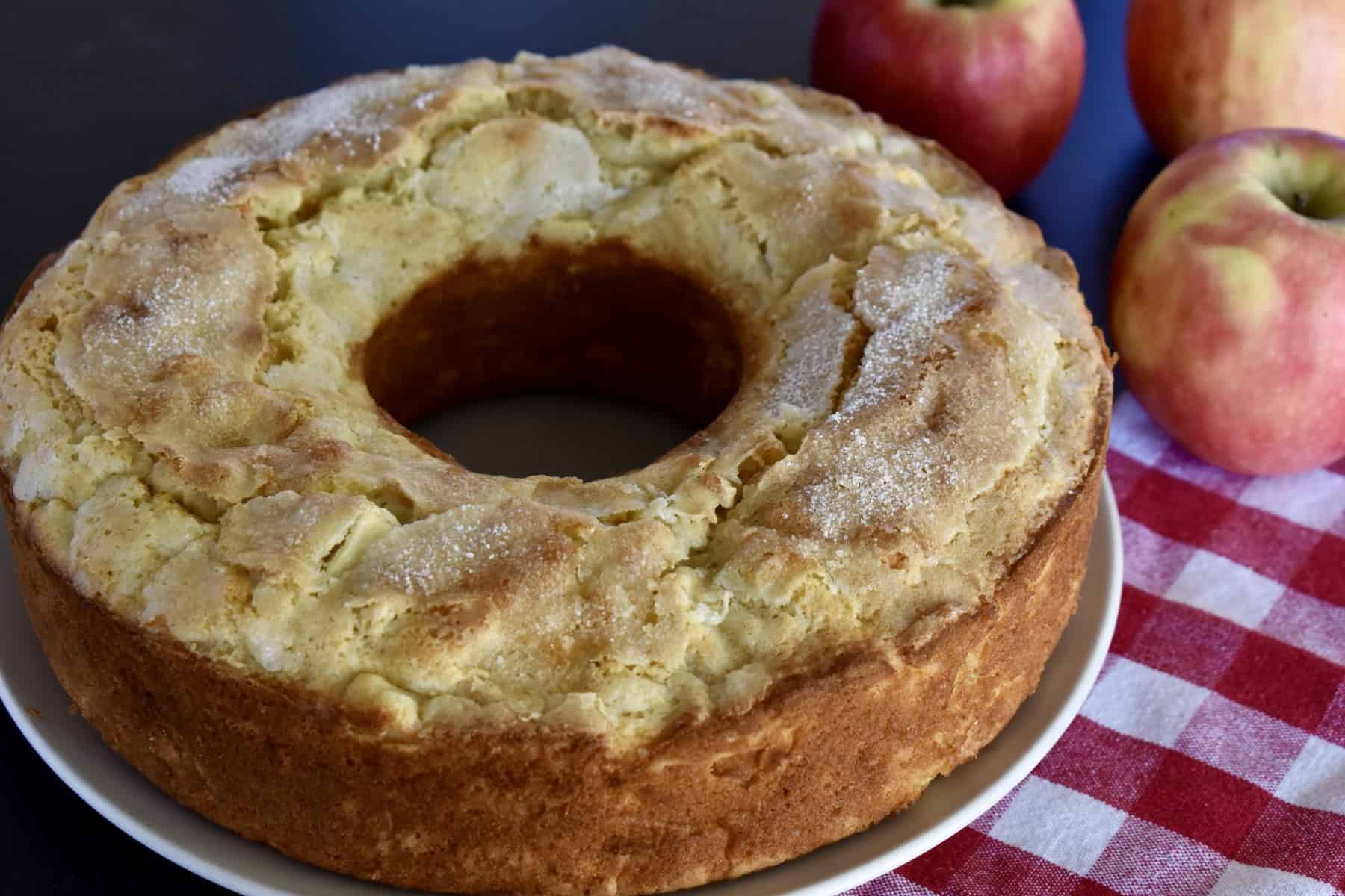Italian Apple Cake on a plate with a checkered cloth and apples in the background. 