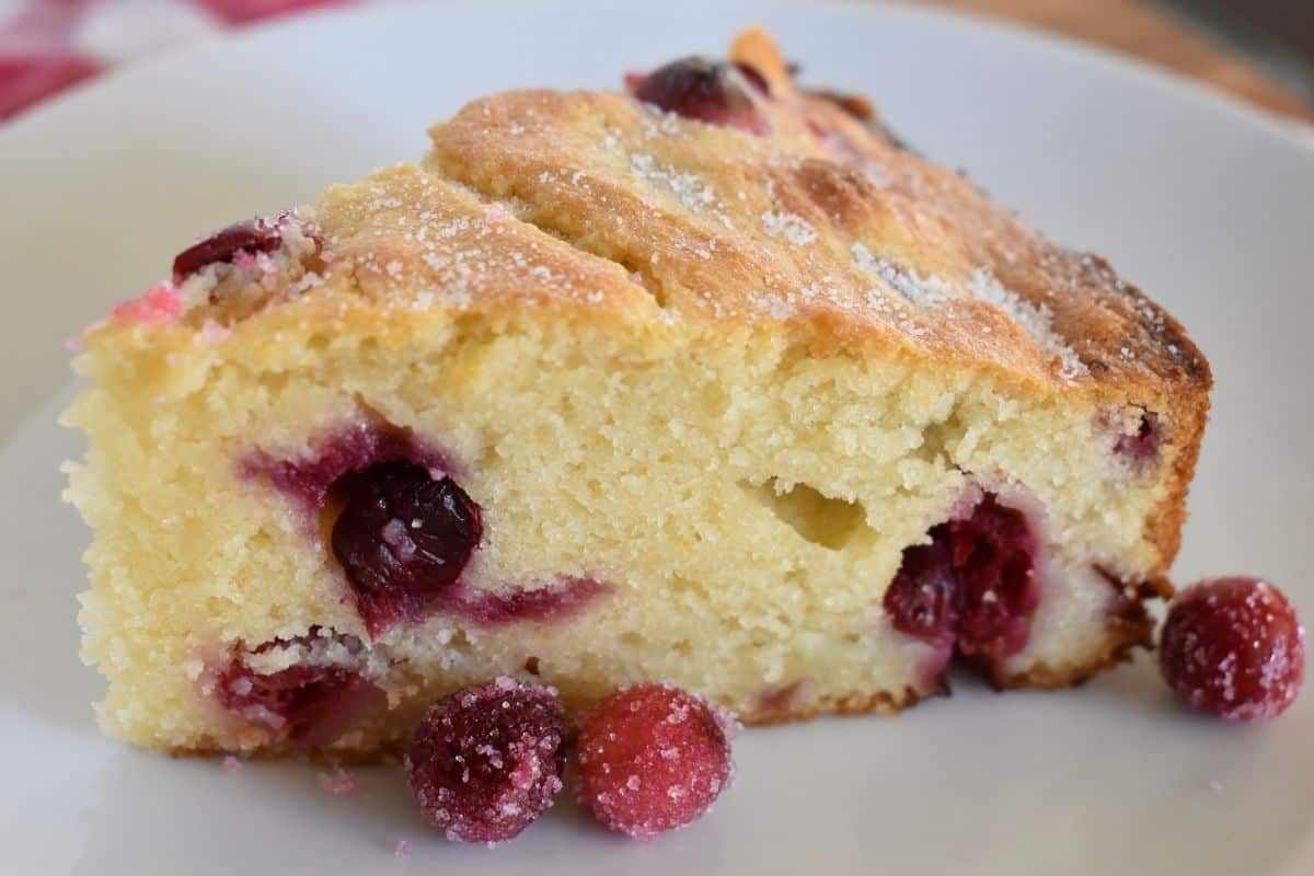 slice of cake on a plate with sugared cranberries next to it. 