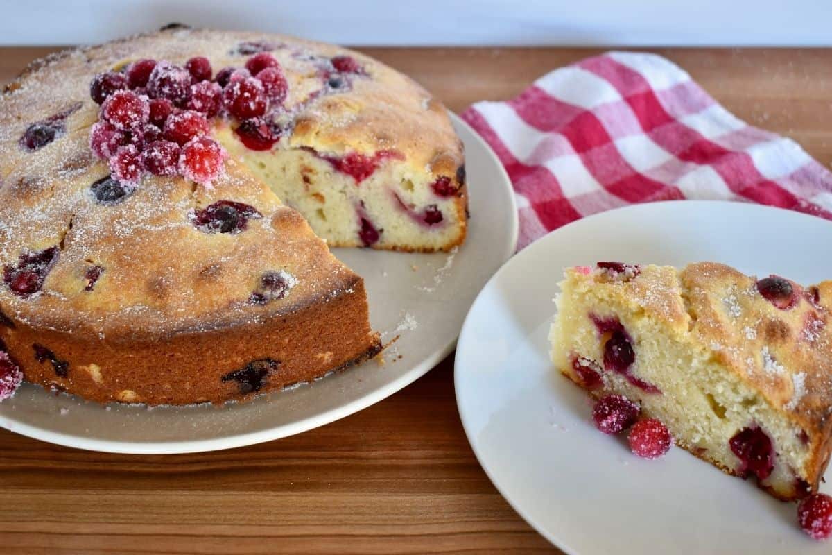 Cranberry ricotta cake on a plate with sugared cranberries on top and a slice of cake on a plate next to it. 