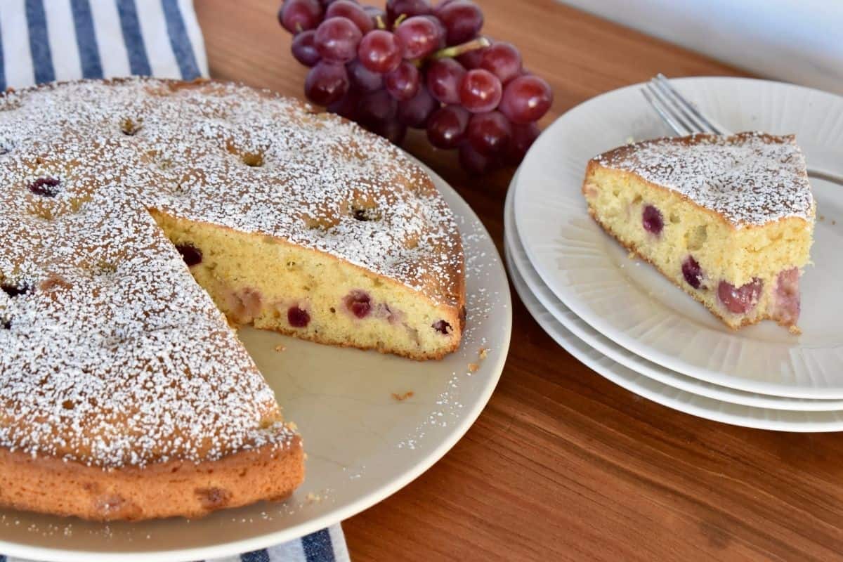 Italian grape cake on a white plate with a slice of cake on white plate and a bunch of grapes. 