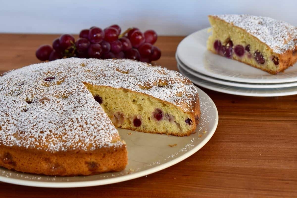 Italian grape cake on a white plate with a slice of the cake and grapes in the background. 