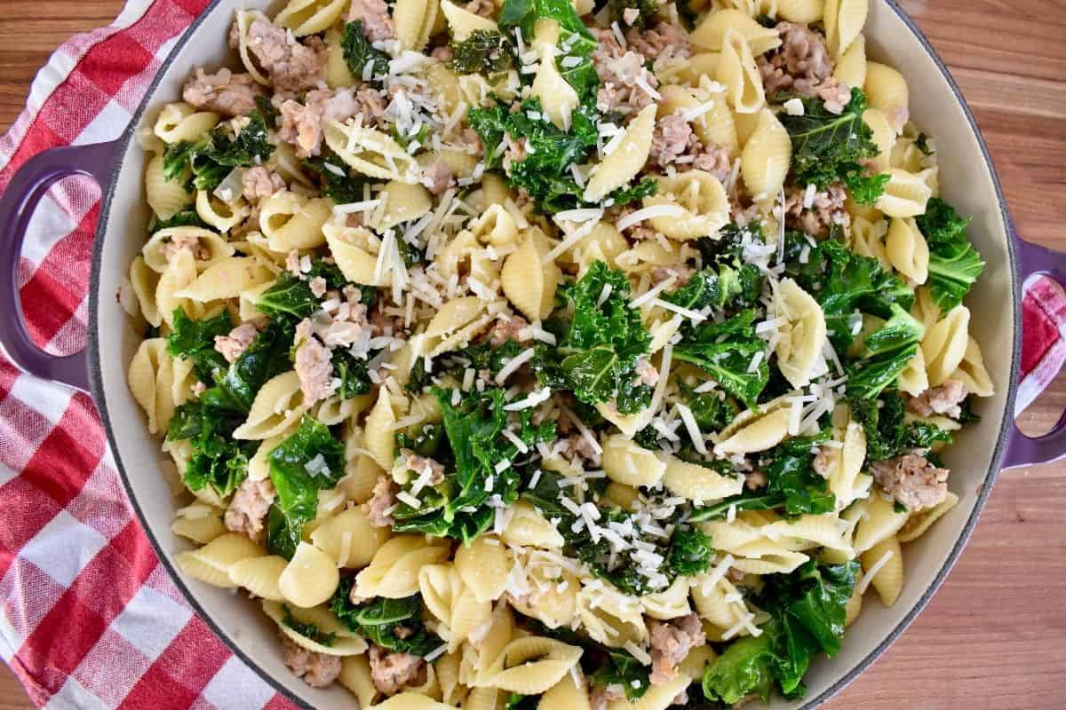 overhead photo of sausage kale pasta in a skillet with a checkered napkin underneath. 