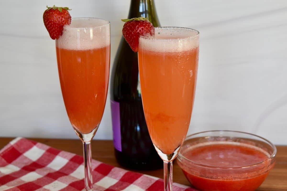 Rossini Cocktail in two champagne glasses on a counter with a bottle of prosecco and strawberry puree. 