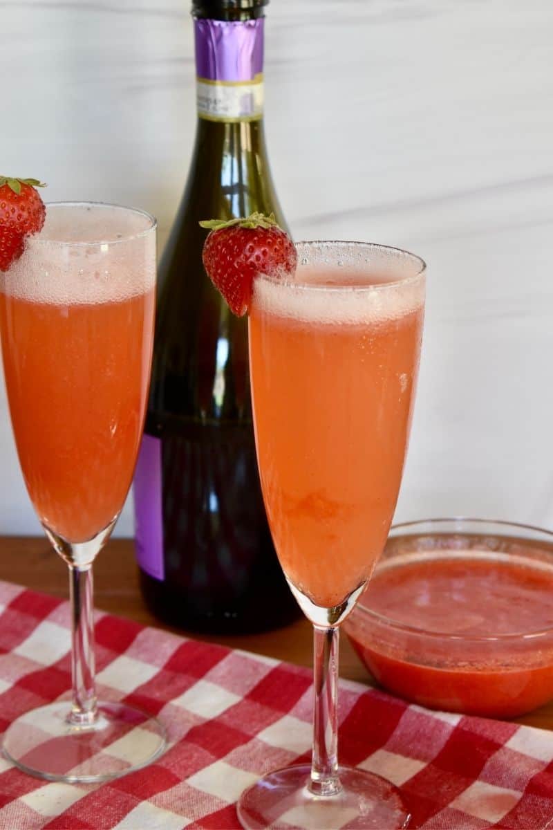 Two glasses of rossini cocktails on a checkered cloth with a bottle of prosecco in the background. 