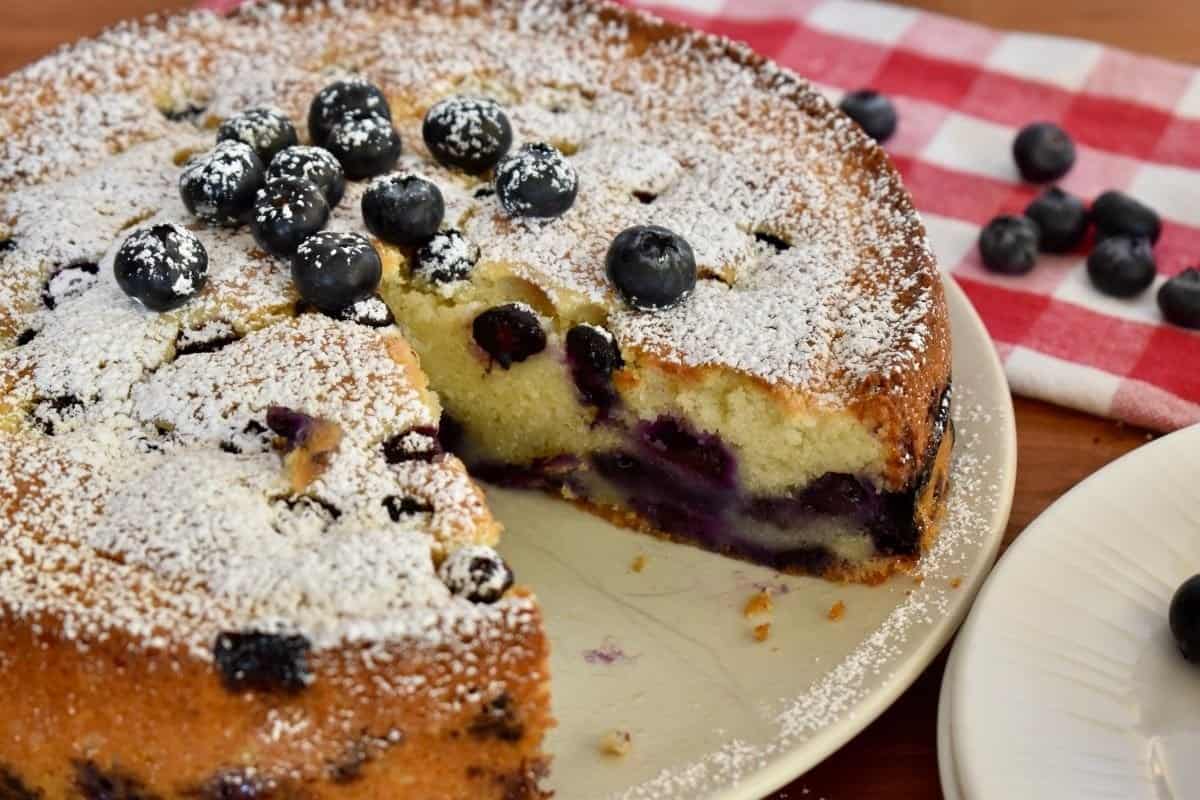 Blueberry Ricotta Cake on a white plate with a slice removed and blueberries in the background. 
