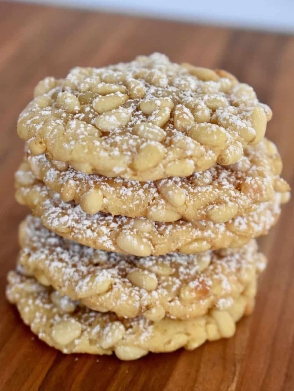 Pignoli Cookies stacked on each other on a wood cutting board. 