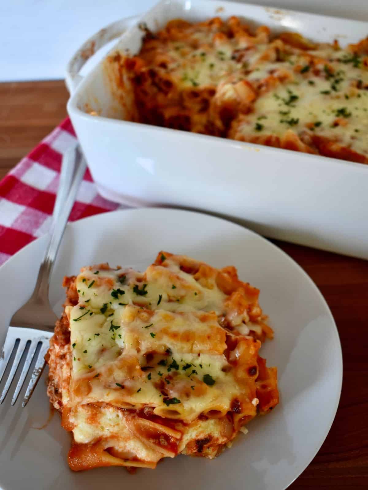 Authentic Italian Mostaccioli recipe with a slice of baked pasta on a plate and the dish of baked pasta in the background. 
