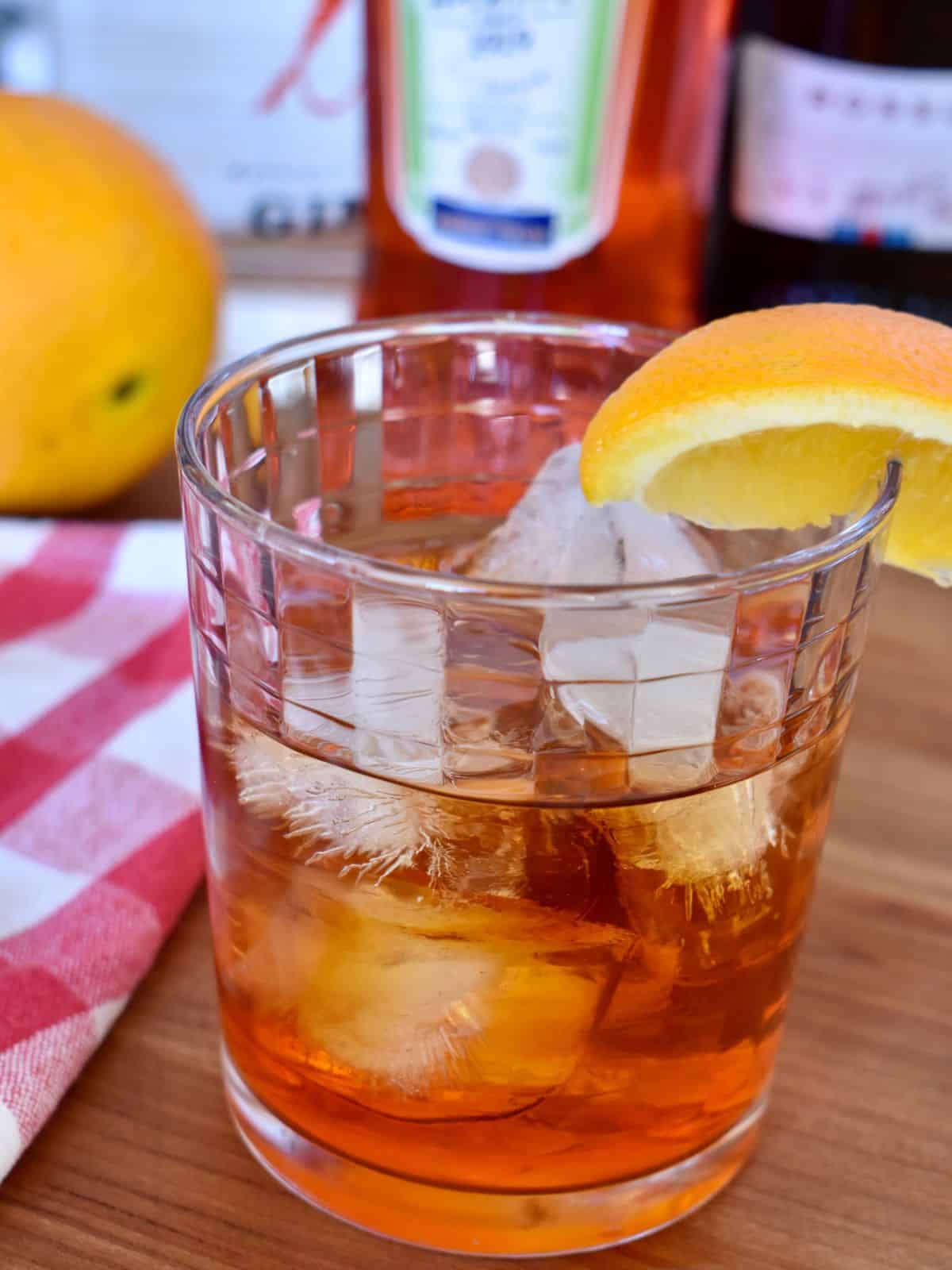 Aperol Negroni on a counter with bottles of gin, sweet vermouth, Aperol, and orange in the background. 