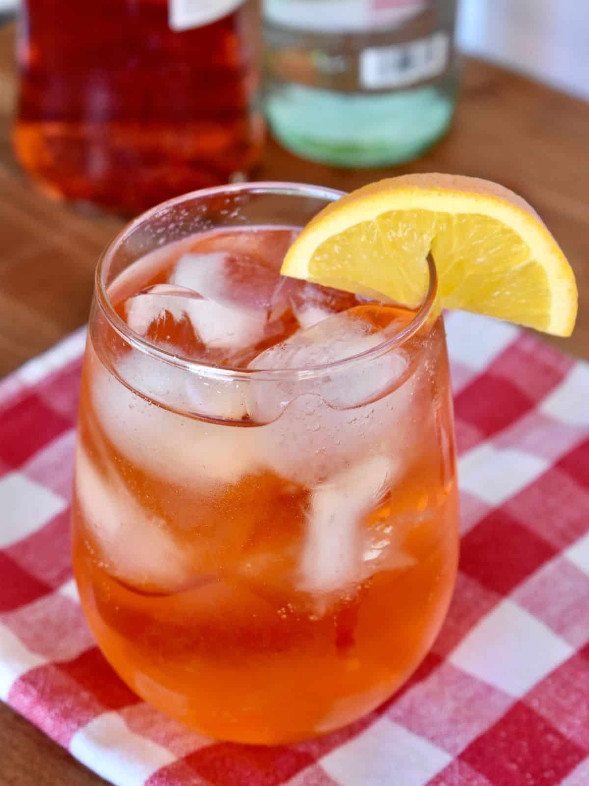 Aperol Soda in a glass with ice and an orange wedge on a checkered napkin. 