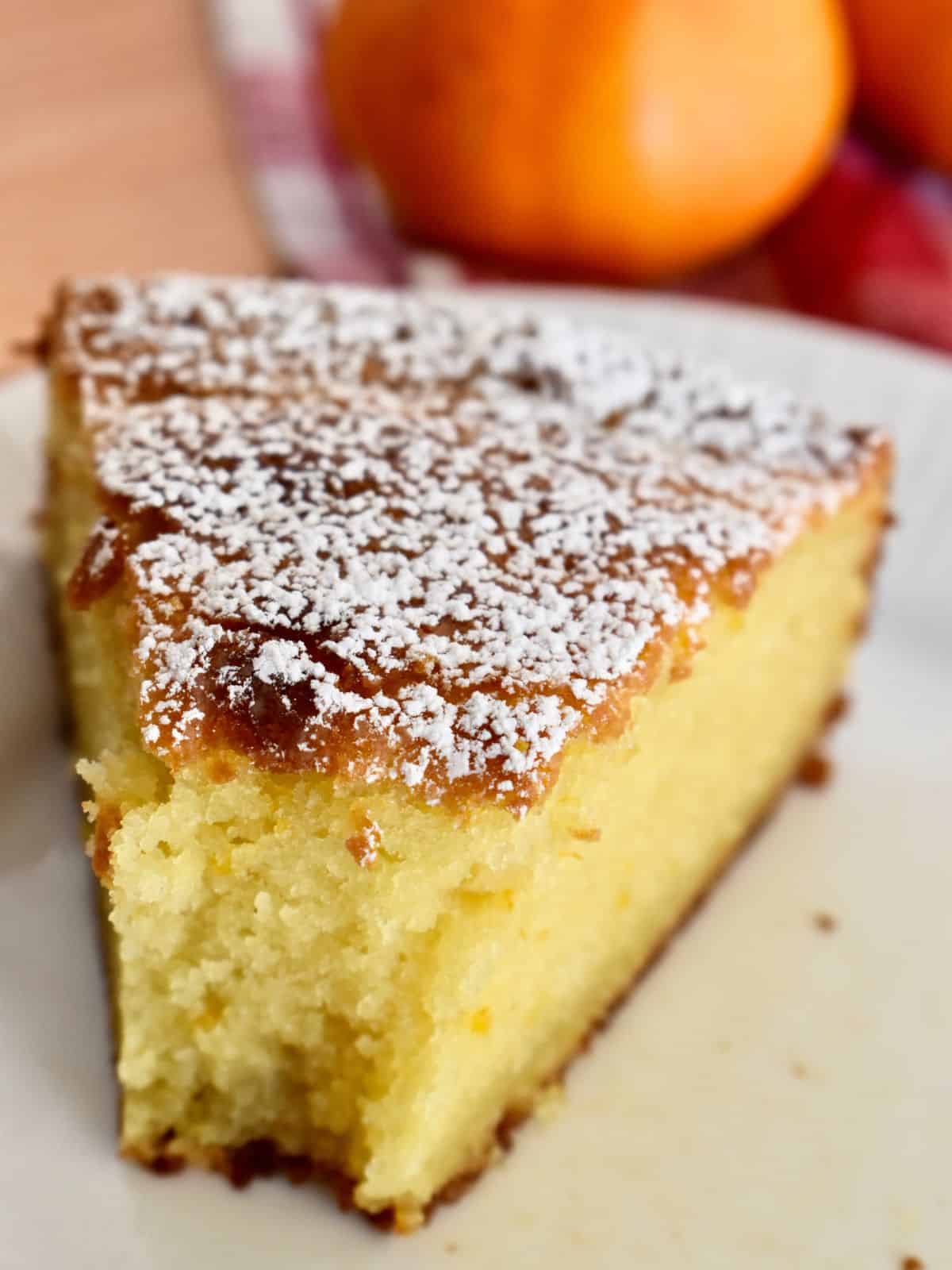 slice of orange ricotta cake on a white plate with a bite taken out of it and an orange in the background. 