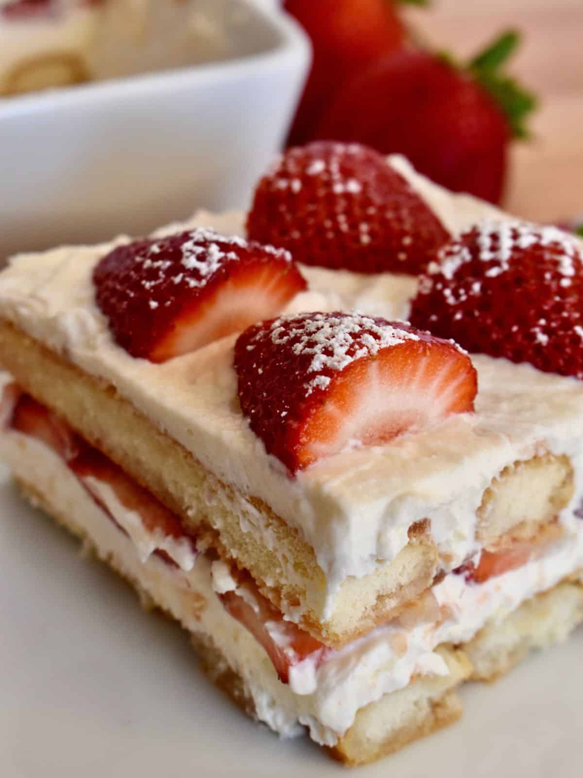Strawberry tiramisu slice on a white plate with strawberries and powdered sugar on top. 