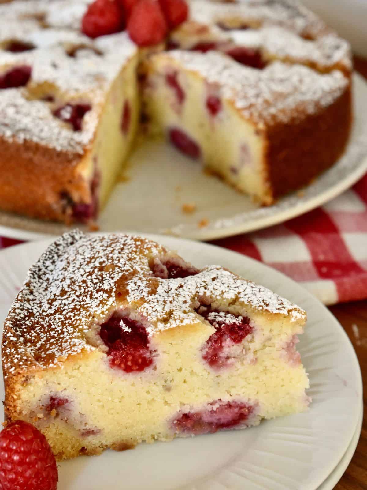 Raspberry Ricotta Cake on a white plate with a cake in the background.