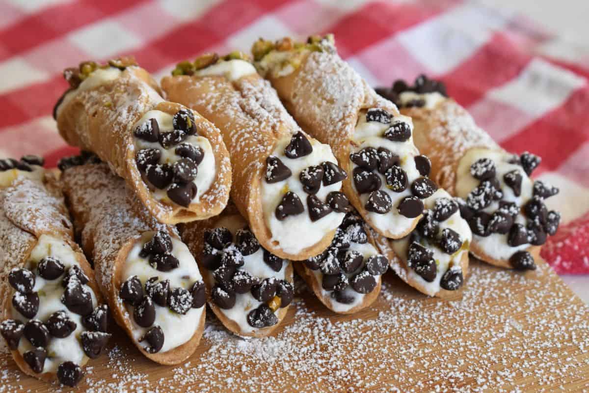 Ricotta Cannolis stacked on each other on a wood board with powdered sugar on top and a checkered napkin in the background.