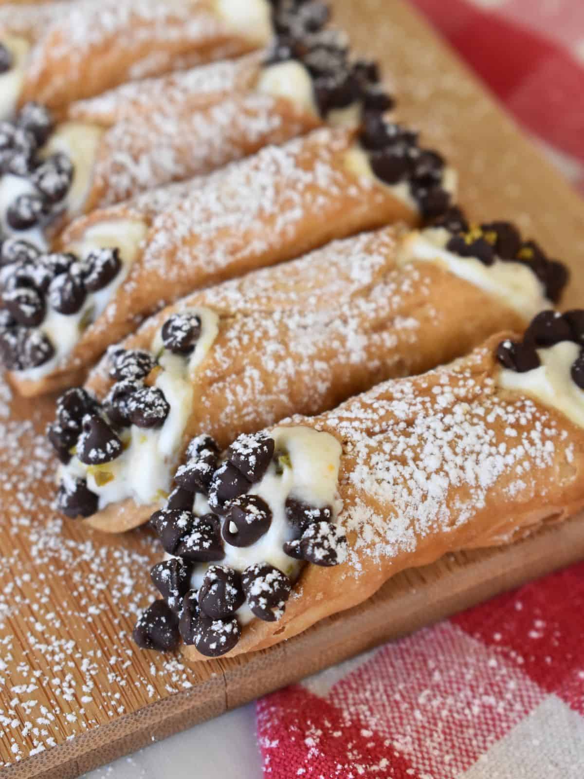 Ricotta Cannolis lined up on a wood board with a checkered napkin underneath.