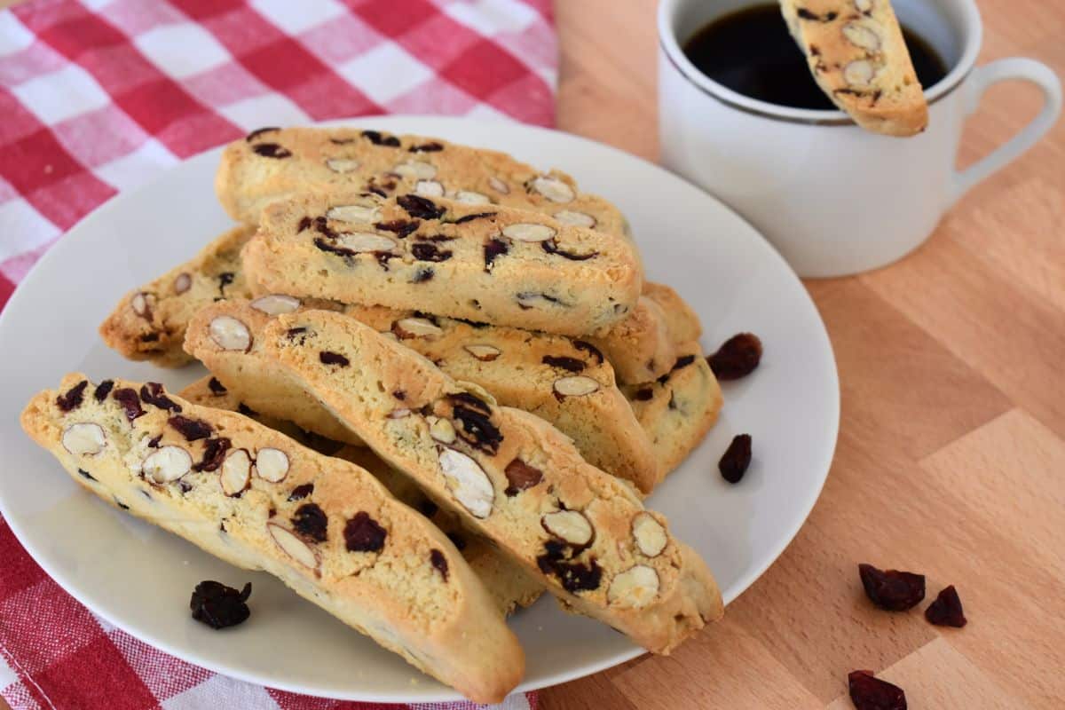 Cranberry Almond Biscotti recipe on a white plate with a cup of coffee and a checkered napkin in the background. 