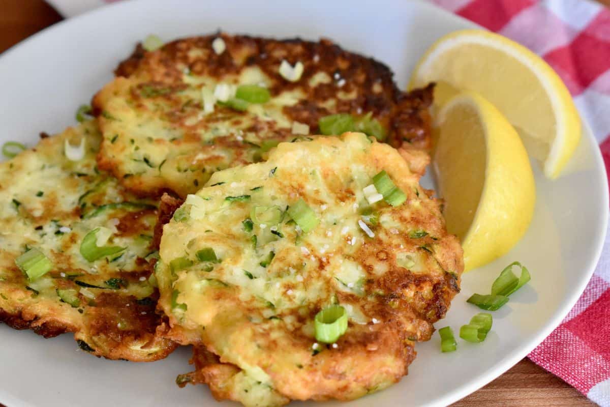 White plate with three zucchini ricotta fritters on it and a slice of lemon. 