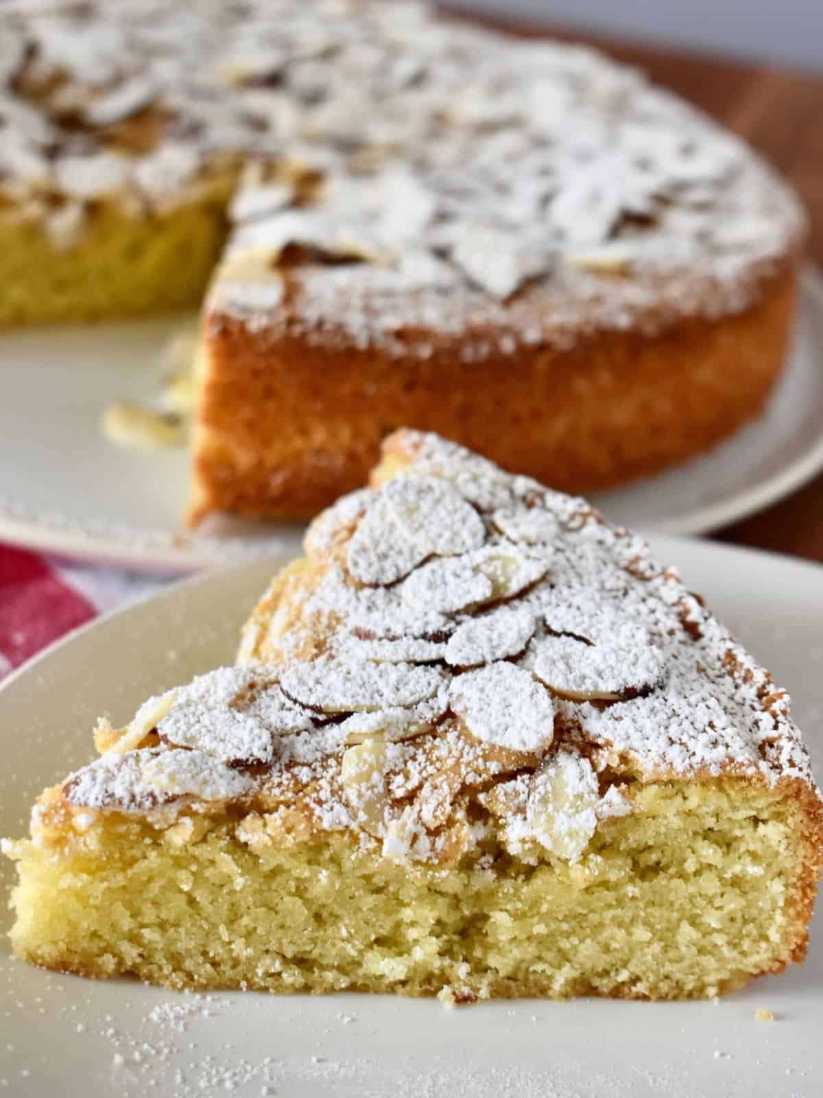 Italian Almond Cake on a white plate with the remaining cake in the background. 