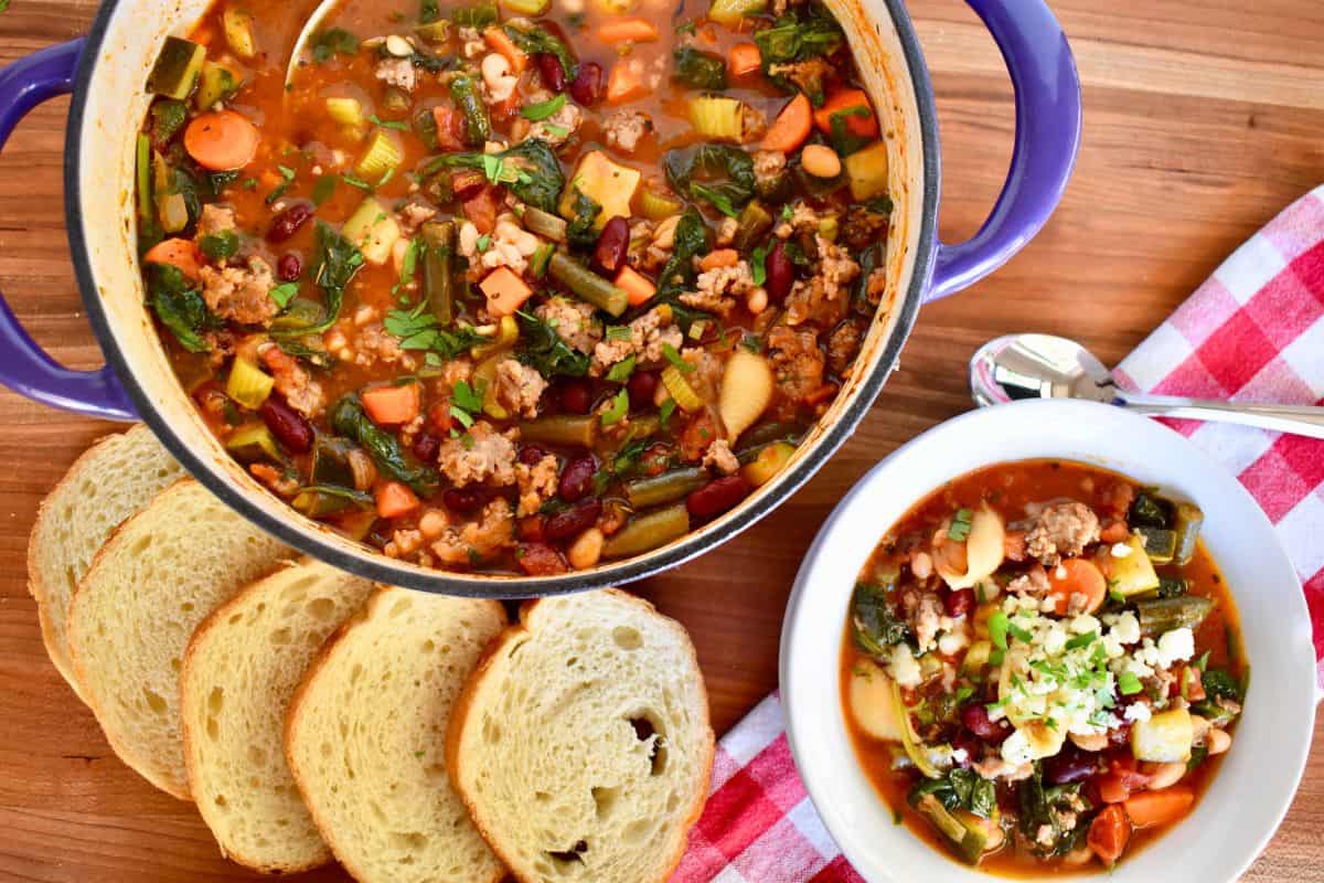 Minestrone soup with sausage in a pot on a wooden board with a bowl of soup and bread. 