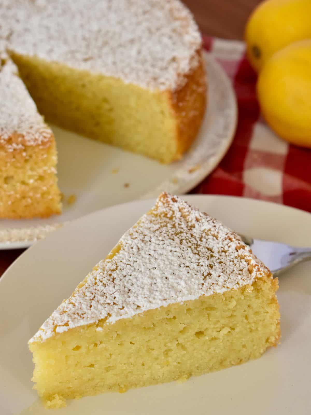 Slice of Italian lemon olive oil cake on a white plate with a fork and lemons in the background. 
