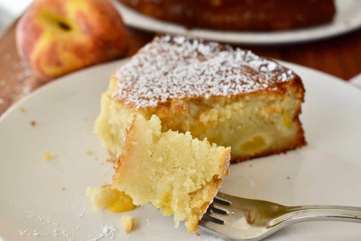 Peach ricotta cake on a white plate with a piece of the cake on a fork and peach in the background. 