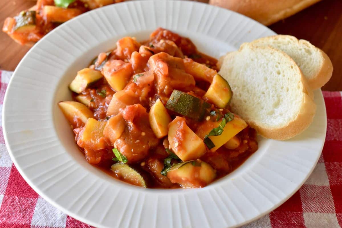 Ciambotta in a white bowl on a checkered napkin with sliced bread. 