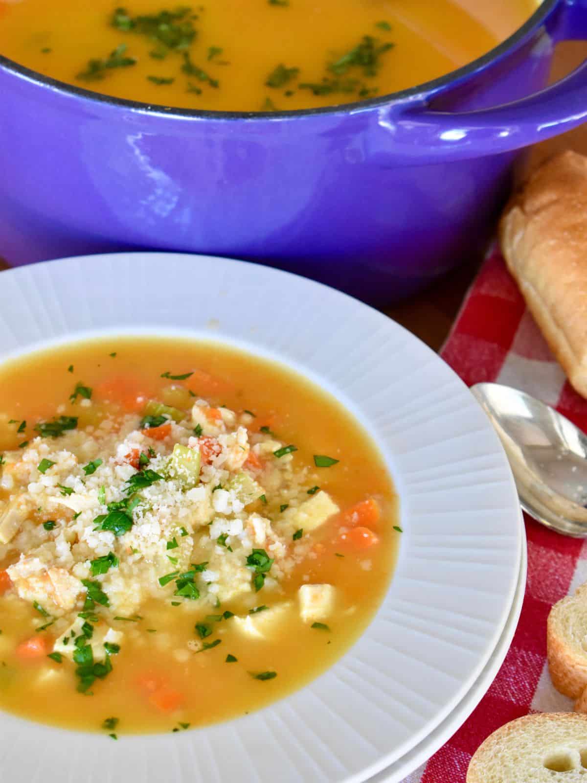 Chicken Pastina Soup in a bowl with a pot of soup behind it. 