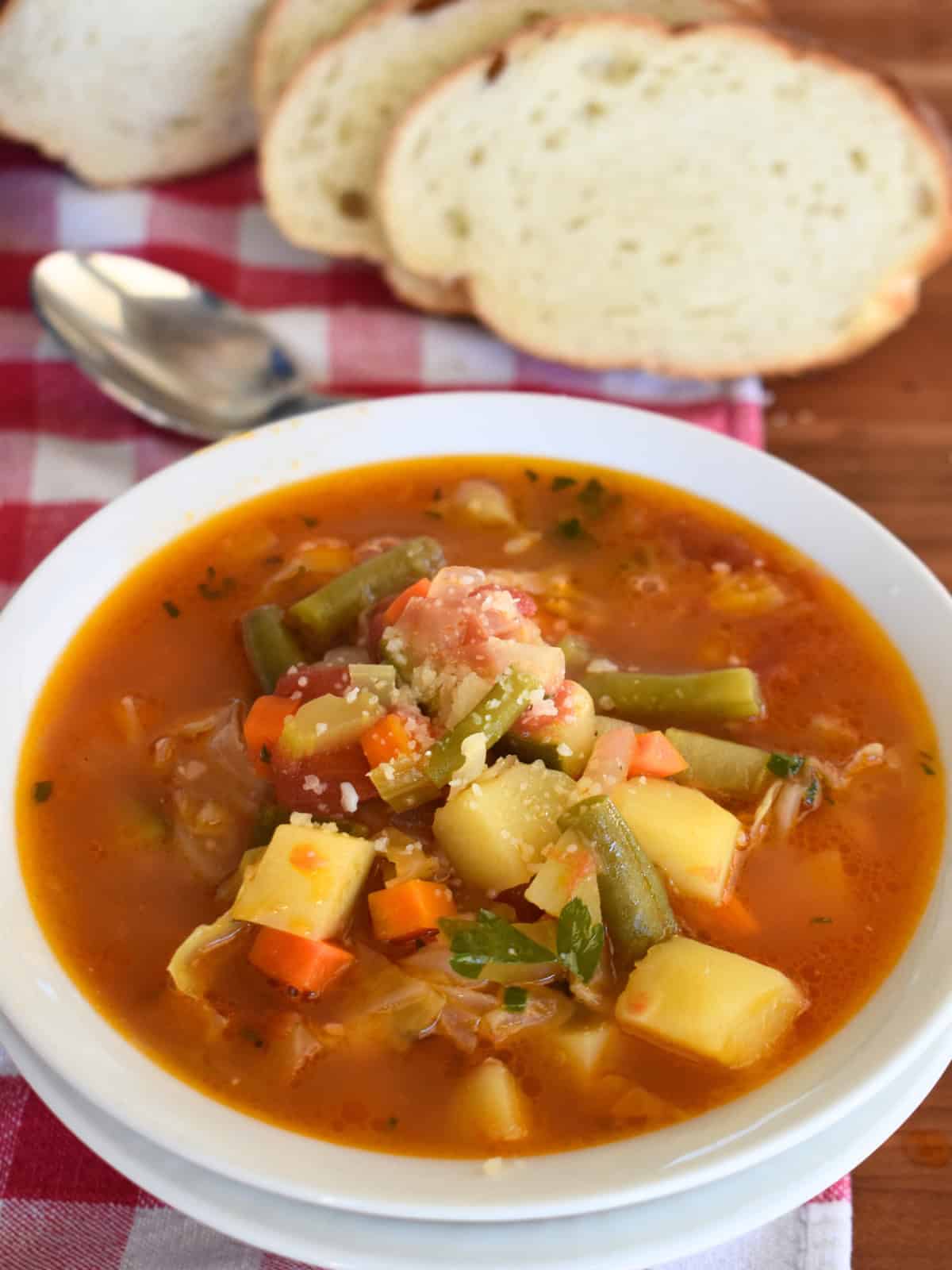 Italian Vegetable Soup in a white bowl with sliced bread in the back. 