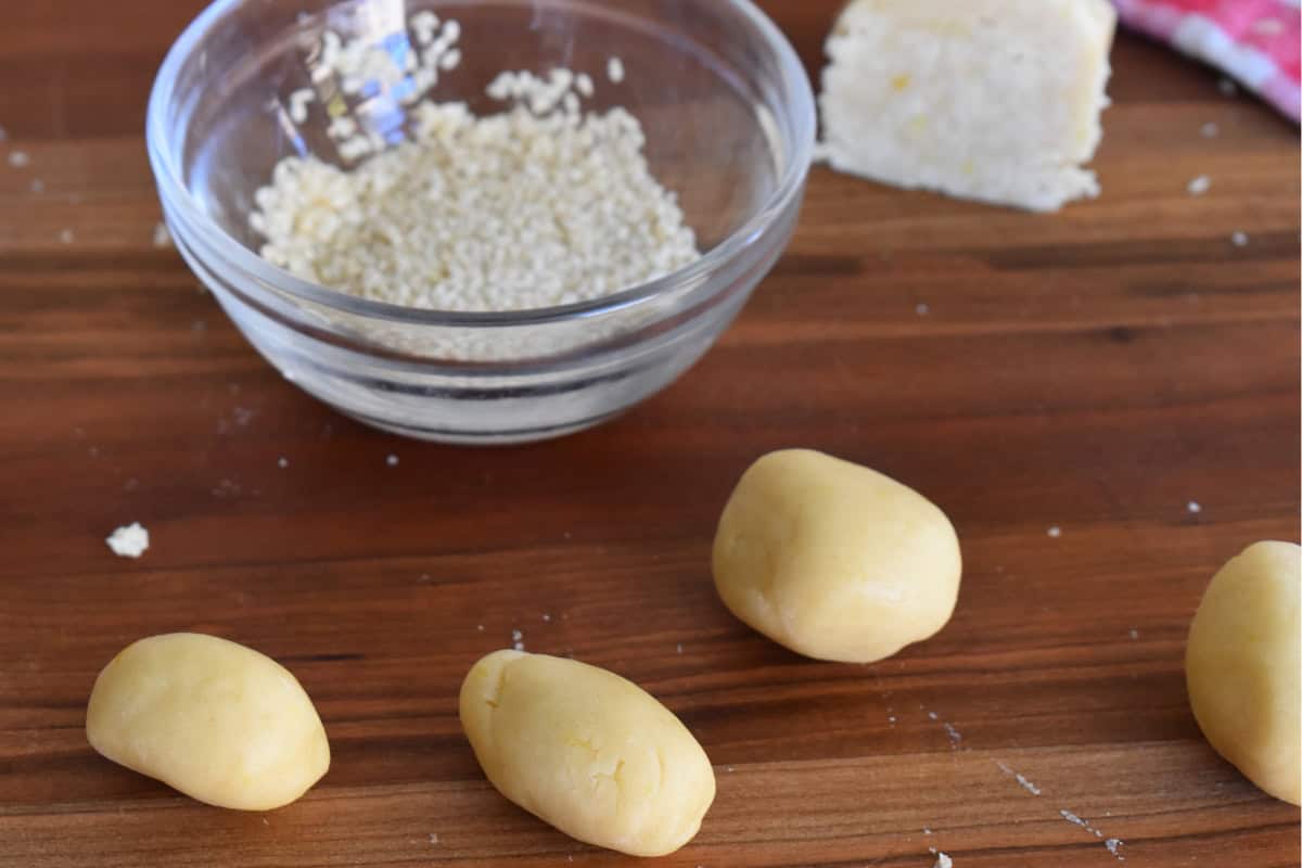 Rounds of cookie dough on a countertop with a bowl of sesame seeds in the background. 