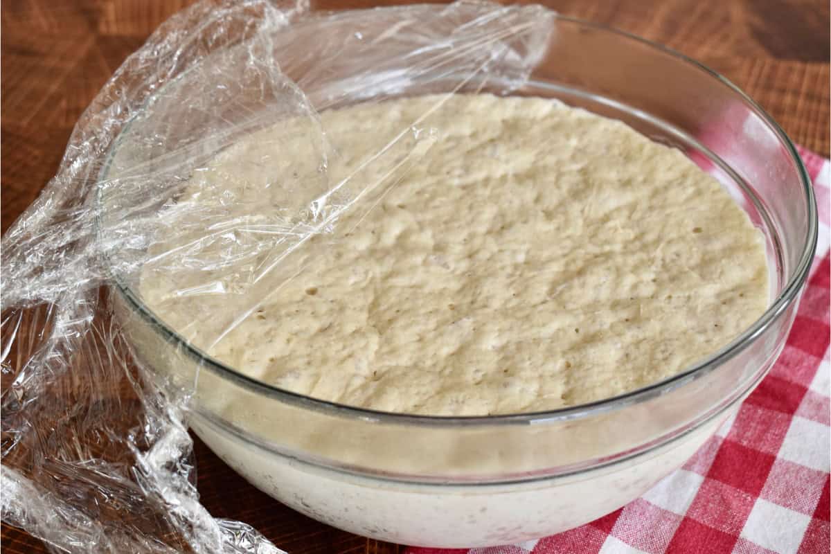 Risen dough in a glass bowl with plastic wrap on top.