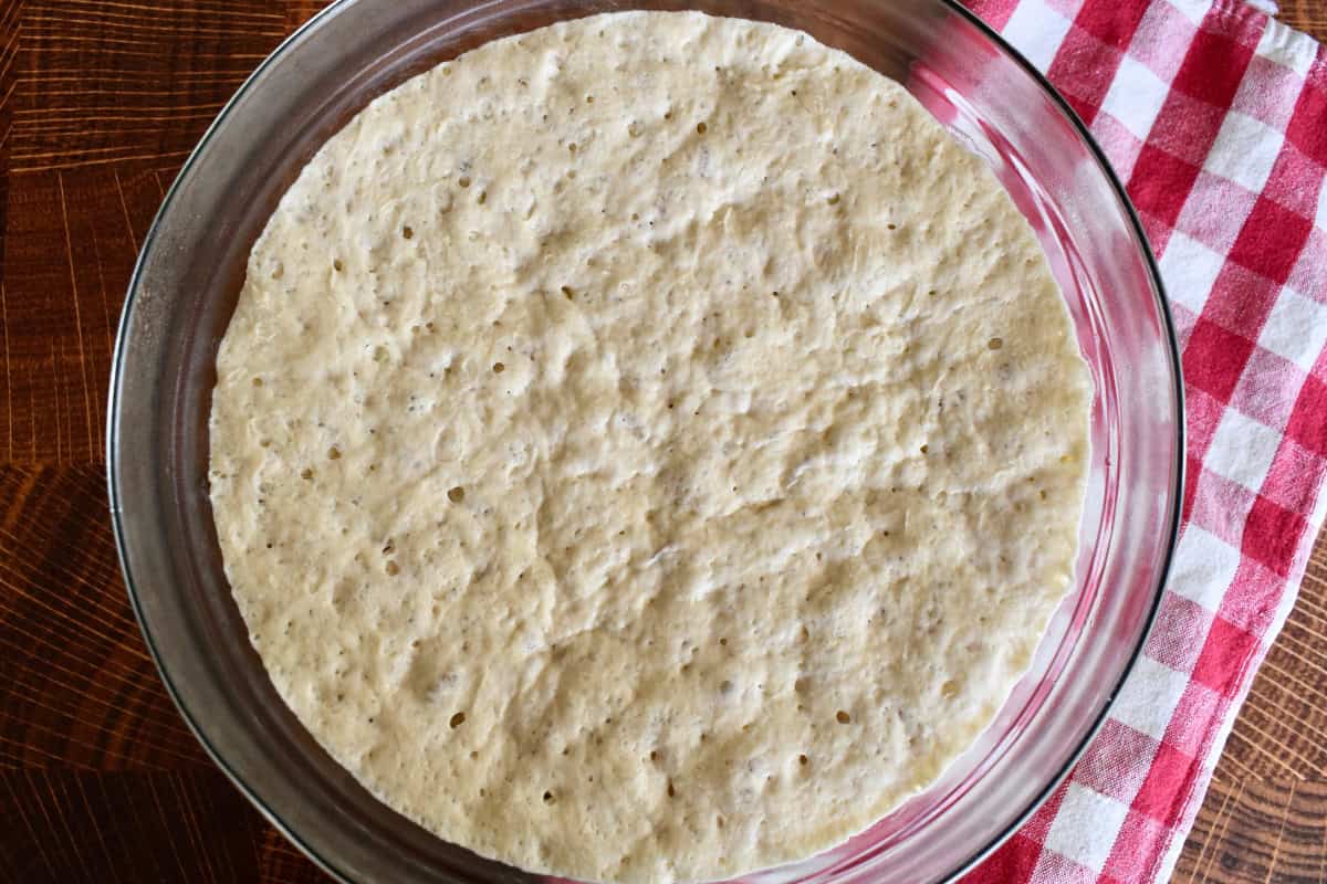 Overnight Pizza Dough in a glass bowl with a checkered napkin underneath. 
