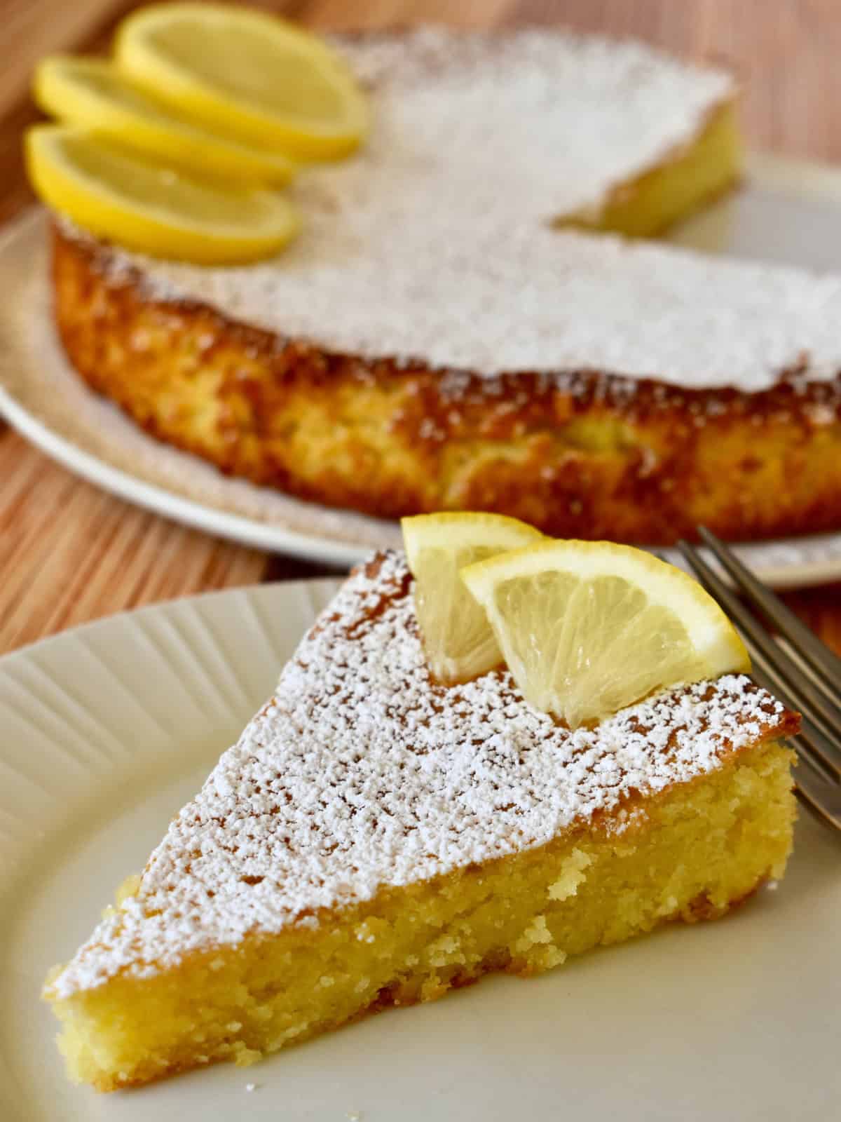 Slice of lemon torta caprese on a white plate with the rest of the cake in the background.