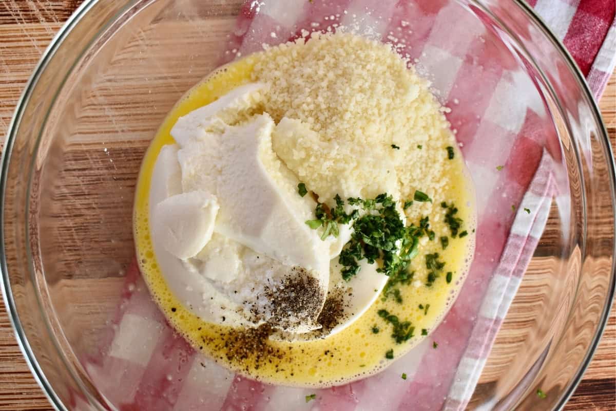 Ingredients in a glass bowl including eggs, ricotta, cheese, and basil. 