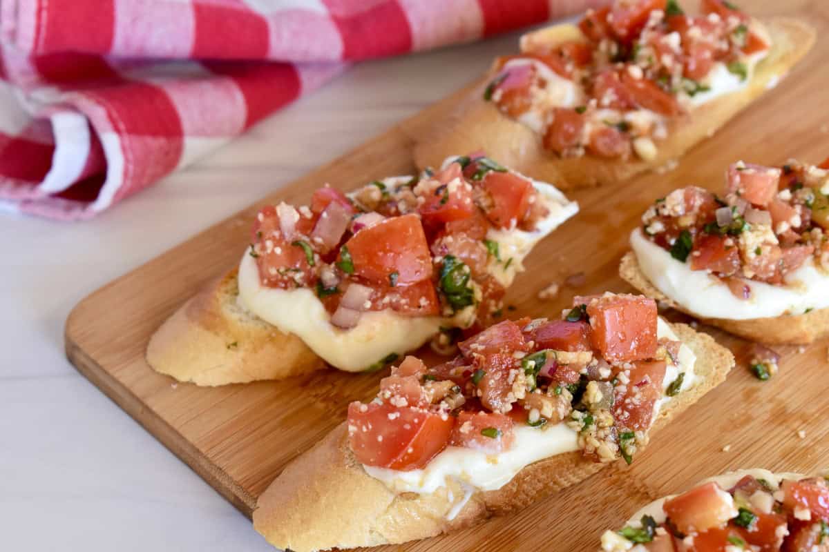 Ricotta Bruschetta on a wood board with a checkered napkin behind it. 