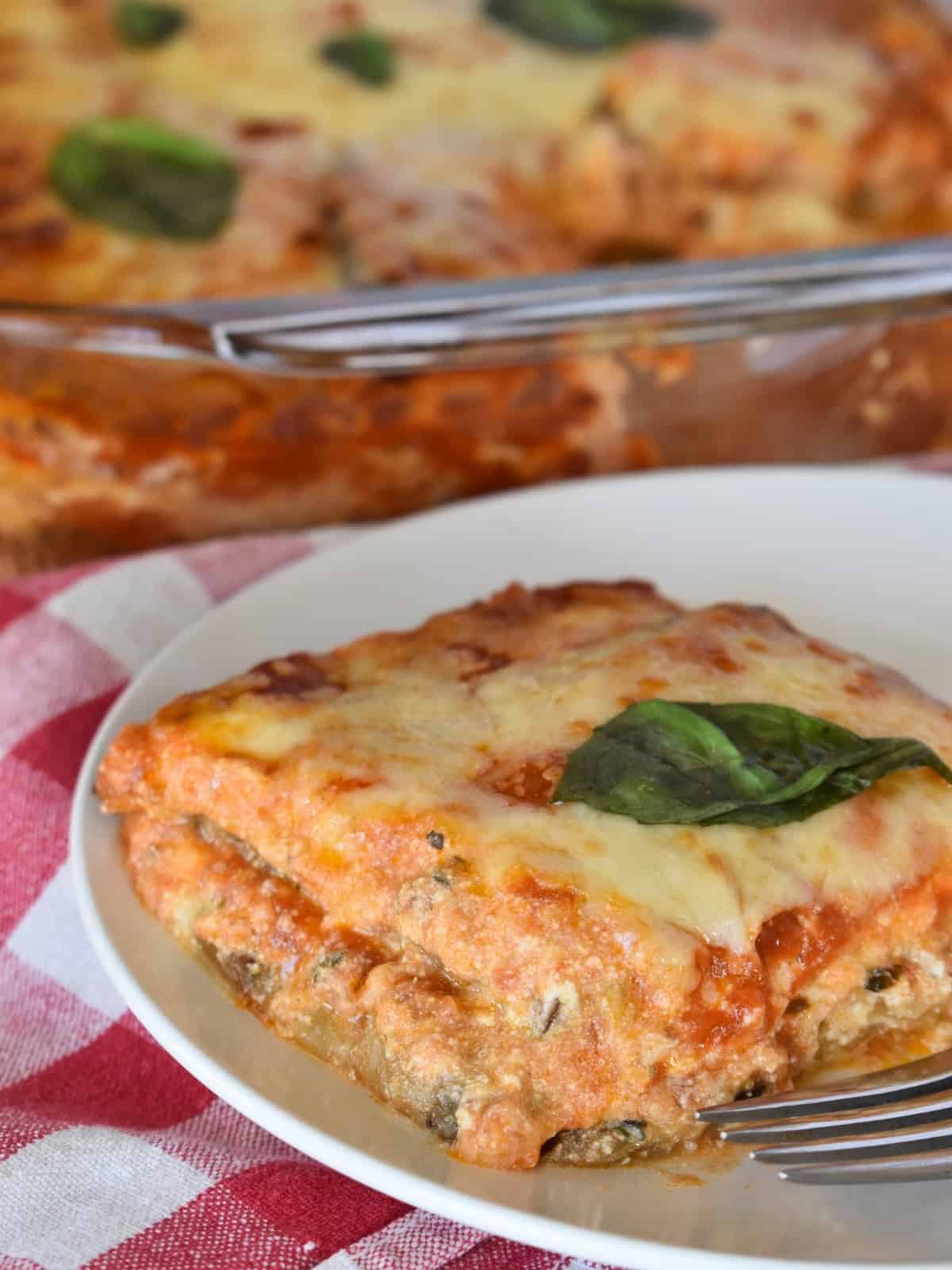 Eggplant lasagna on a white plate with a pan of it in the background. 