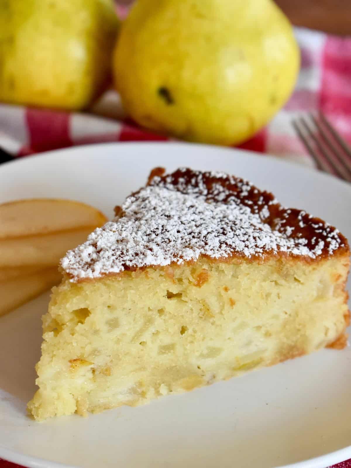 Slice of pear ricotta cake on a white plate with pears in the background. 